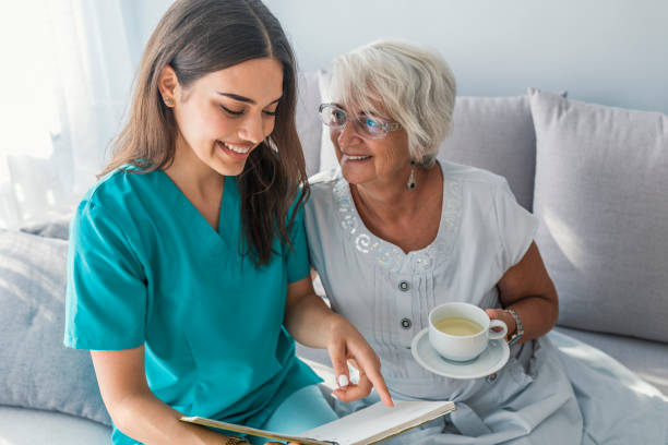 Happy elder woman sitting on white sofa and listening to nurse reading a book out loud. Professional helpful caregiver comforting smiling senior woman at nursing home